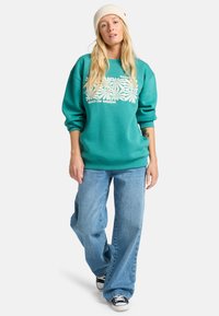 Felpa teal con grafica floreale bianca, vestibilità ampia, polsini a coste, abbinata a jeans leggeri a vita alta in denim blu chiaro e sneakers nere.