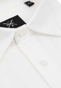 Biała koszula z kołnierzykiem, o gładkiej fakturze materiału, z przodem zapinanym na guziki oraz czarną metką marki przy kołnierzu.