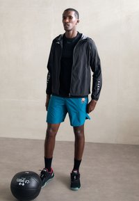 Sort letvægtsjakke med lynlås, teal shorts og sorte sko med røde detaljer. Har et sømløst design og atletisk pasform.