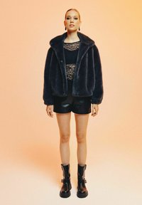 Veste en fausse fourrure de couleur bleu marine foncé, avec un large col et une fermeture à boutons, associée à un top en dentelle, un short noir texturé et des bottes noires imposantes.