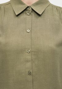 Πουκάμισο από λινού χρώματος ελιάς με μυτερό γιακά και πέντε κουμπιά σε κατακόρυφη ευθεία στο μπροστινό μέρος.