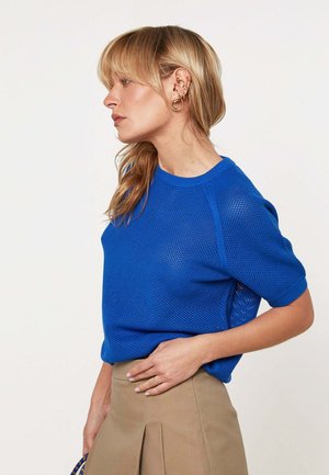 Blonde vrouw in een blauw gebreide korte mouw top en beige rok, een blauwe handtas vasthoudend, kijkt naar links tegen een neutrale achtergrond.