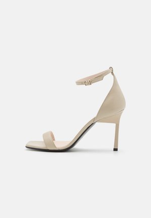Beige leather high-heeled sandal with thin ankle strap, open toe, and a sleek, tapered silhouette. Smooth texture and minimalistic design.