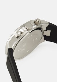 Ruostumattomasta teräksestä valmistettu kellokotelo, jossa on kolme hopeista painiketta ja musta silikonihihna. Kiiltävä ja mattapintainen lopetus, joka on sileä ja heijastava.