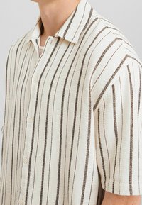 Man i en kortärmad krämfärgad skjorta med smala vertikala mörkbruna ränder och en vit t-shirt synlig vid kragen.