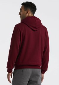 Bordeaux hoodie met een trekkoordcapuchon, lange mouwen en geribbelde manchetten. Soepele textuur en casual pasvorm. Gedragen over grijze broeken.