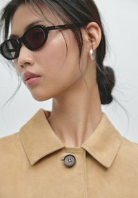 Vrouw met donker haar in een knot, draagt zwarte ovale zonnebril, zilveren oorbellen en een beige gekraagde jas met een knoopsluiting.