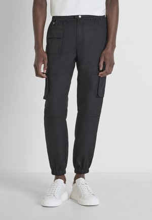 Zwarte cargobroek met een elastische tailleband, zijzakken, taps toelopende pijpen en elastische boorden, gecombineerd met witte sneakers op een lichte achtergrond.