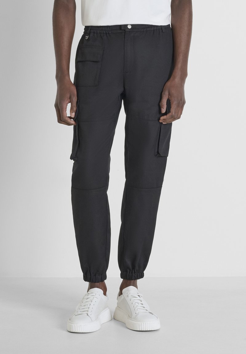 Zwarte cargobroek met een elastische tailleband, zijzakken, taps toelopende pijpen en elastische boorden, gecombineerd met witte sneakers op een lichte achtergrond.