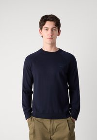 Mornarska pulover s krojem čoln iz mehkega pletenega blaga, z raglan rokavi in diskretno logotipno podrobnostjo na prsih. V kombinaciji s kaki hlačami.