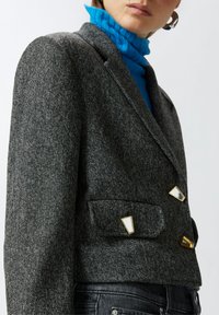 Sivi teksturirani blejzer s zlatnim i bijelim trokutastim naglascima na džepovima, krojenog kroja i reverima, nosi se preko plave rolke.