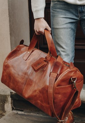 Brown leather duffel bag with structured shape, dual handles, adjustable shoulder strap, and a tag attached featuring "Holatier."