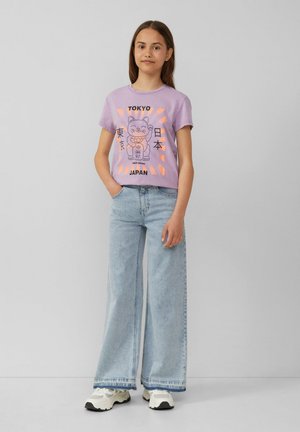 T-shirt lilas avec un design graphique d'un chat et le texte Tokyo. Associé à un jean large bleu clair et des baskets blanches.