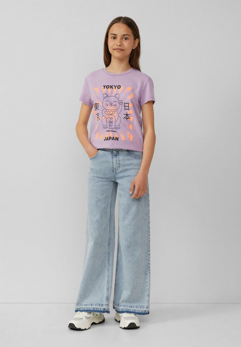 Lila T-shirt met een grafisch ontwerp van een kat en de tekst 'Tokyo'. Gepaarde met lichtblauwe wijdere jeans en witte sneakers.