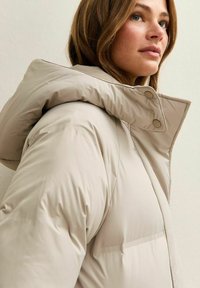 Chaqueta de invierno acolchada beige con cuello alto, detalles de botones y un diseño elegante y aerodinámico. Fabricada con un material suave e impermeable.