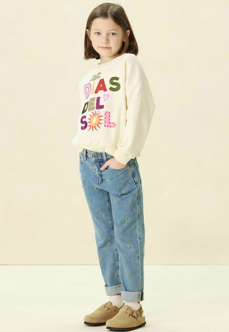 Sweatshirt de couleur claire avec le design "DÍAS DEL SOL", jeans bleus avec de petits motifs de cœurs jaunes, et sabots beiges.