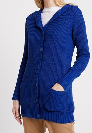 Blauwe gebreide cardigan met knopen en voorzakken, gedragen over een wit shirt en beige broek.