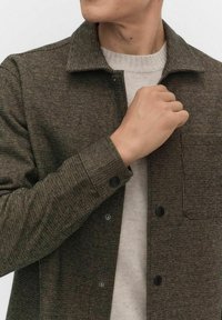 Bruin geruit jack met een kraagontwerp, knoopsluitingen en een borstzak, gelaagd over een lichtbeige gebreide trui.