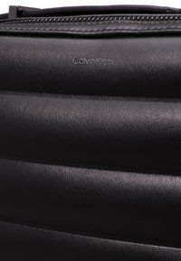 Czarna skórzana torba o teksturowanej powierzchni, z delikatnie wytłoczonym logo Calvin Klein i zamknięciem na zamek błyskawiczny u góry.