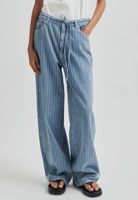 Világoskék farmer, széles lábú nadrág függőleges fehér csíkokkal, állítható derékrésszel és oldalapatokkal. Az varrások látható varrási részleteket tartalmaznak.