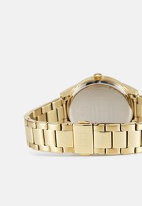 Orologio in metallo dorato con un design a bracciale a maglie. Presenta un quadrante rotondo, una corona sul lato e una chiusura con dettaglio del logo inciso.