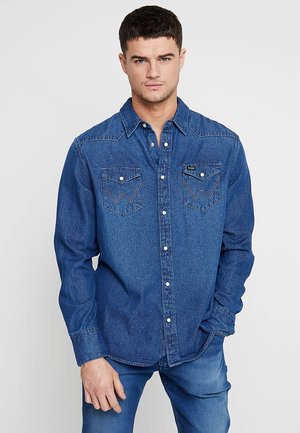 Camisa de mezclilla con un diseño clásico de botones, dos bolsillos en el pecho con detalles de costura y un color azul medio. Mangas largas y cuello puntiagudo.