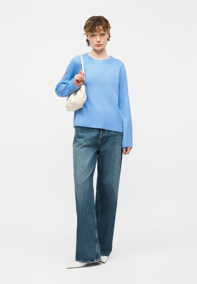 Νεαρό άτομο που φορά ένα ανοιχτό μπλε πουλόβερ, φαρδιά τζιν παντελόνια, λευκά μυτερά παπούτσια και κρατά μια μικρή λευκή τσάντα ώμου.