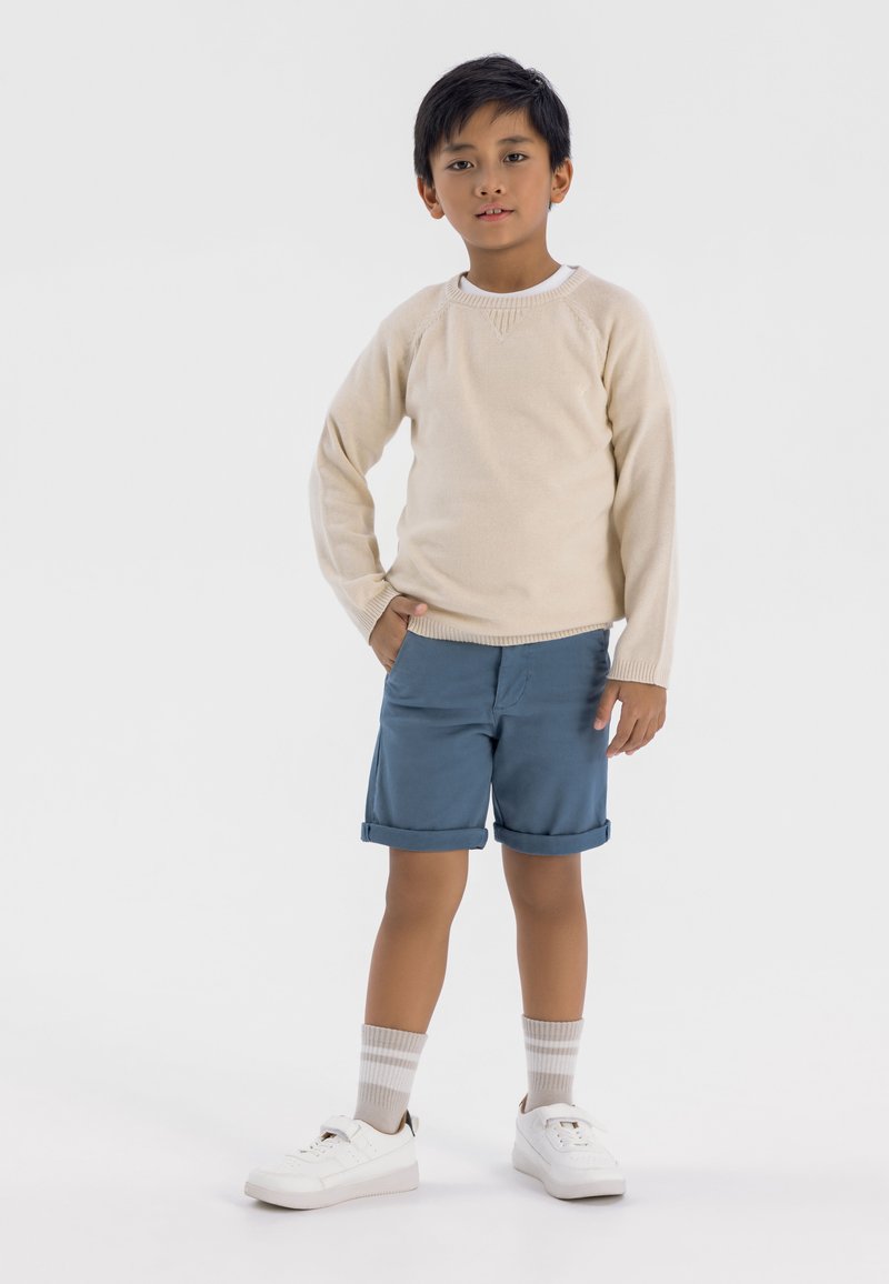 Pull en tricot beige, shorts bleus retroussés, baskets blanches et chaussettes rayées. La tenue présente un style classique avec une coupe décontractée.