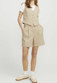 Beige sleeveless vest with buttons paired with high-waisted beige shorts. Model wears white t-shirt and sneakers, showcasing a casual look.