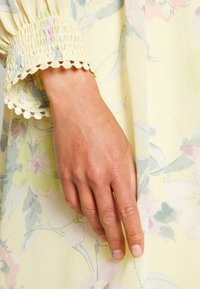 Detalhe de vestido floral com tecido amarelo, apresentando cores pastel suaves, mangas frufru texturizadas com acabamento em renda, e uma silhueta suave e fluida.