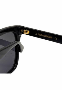 Svarta solglasögon med en blank finish, utrustade med grå linser och guldaccent på skalmen, graverad med "Soho Copenhagen."