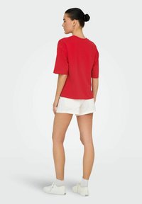 Rode katoenen t-shirt met korte mouwen; gedragen met witte shorts en witte sneakers. De achterkant toont een relaxte pasvorm en minimale details.