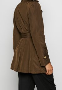 Person klädd i en brun trenchcoat med bälte, sedd från ryggen, med svarta byxor synliga nedanför kappan.