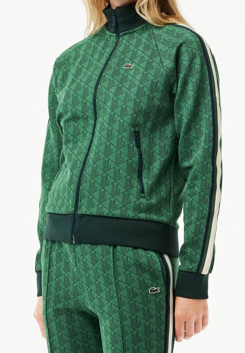 Personne portant un survêtement zippé vert à motifs avec des bandes blanches et bleu marine sur les manches et le pantalon, avec un petit logo crocodile.