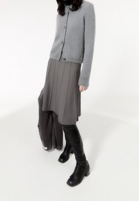 Kvinna i grå kofta och kjol, som håller en mörk jacka och svarta knähöga stövlar, står mot en vit bakgrund.