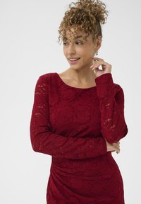 Abito rosso in pizzo con maniche lunghe, caratterizzato da un colletto rotondo e dettagli arricciati ai lati. Il tessuto presenta un motivo floreale e una finitura texturizzata.