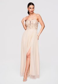 Femme portant une robe de soirée beige sans manches avec un corsage en sequins, un décolleté en V profond, une fente haute jusqu'à la cuisse et des talons argentés ouverts, posant devant un fond uni.