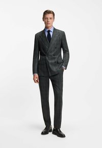 Completo doppiopetto grigio scuro con un motivo a quadri, abbinato a una camicia azzurro chiaro, cravatta blu navy e scarpe eleganti nere.