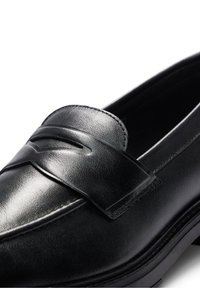 Mocassin en cuir noir avec une bride à pièce de monnaie et une finition lisse, présenté en gros plan sous un angle sur un fond blanc.