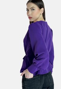 Blouse en satin violet avec des manches longues bouffantes et un détail froncé à la taille, associée à un jean noir. Le tissu a une texture lisse et brillante.