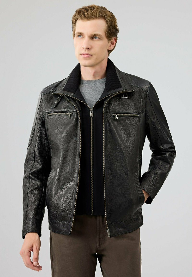 Zwarte leren jas met een gestructureerde afwerking, een hoge kraag en twee ritszakken aan de voorkant. Draag over een grijze shirt. Bruine broek zichtbaar.