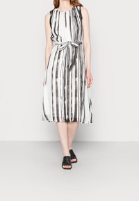 Ärmelloses Midikleid in Schwarz und Weiß mit vertikalen Streifen; der Stoff hat eine glatte Textur; enthält ein gebundenes Taillendetail; kombiniert mit schwarzen Mules.