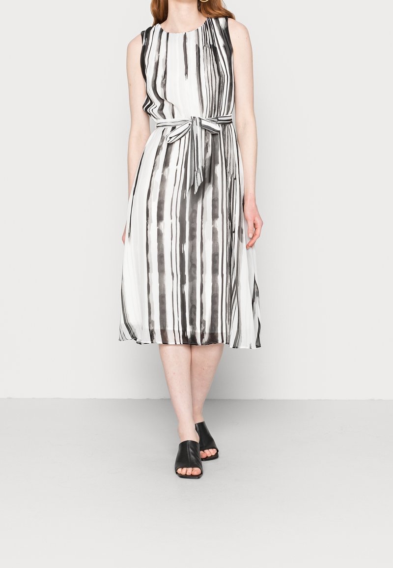 Ärmelloses Midikleid in Schwarz und Weiß mit vertikalen Streifen; der Stoff hat eine glatte Textur; enthält ein gebundenes Taillendetail; kombiniert mit schwarzen Mules.