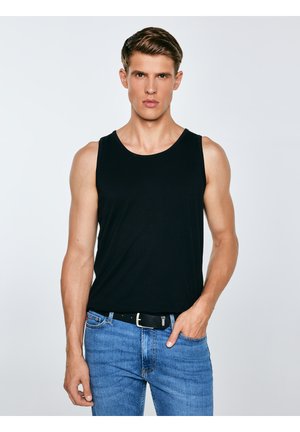 Jauns vīrietis melnā krekliņā bez piedurknēm un zilās džinsās, stāvot ar vienu roku kabatā pret vienkrāsainu gaišu fonu.