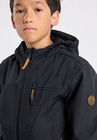 Musta pehmeäkuorinen takki, jossa on vetoketjukiinnitys, huppu ja kontrastiset kultaiset yksityiskohdat. Takissa on pyöreä logopatch ja edessä vetoketjulliset taskut.