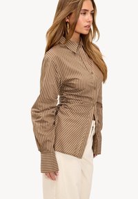 Brown striped button-up shirt with long sleeves, ruched detail at the sides, collared neck, and smooth texture. Paired with light-colored pants.