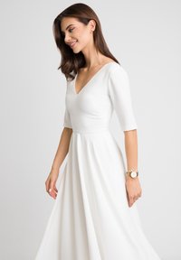 Biała sukienka z dekoltem w kształcie litery V i krótkimi rękawami. Luźna spódnica, gładka faktura. Złoty zegarek na nadgarstku. Prosty i elegancki design.