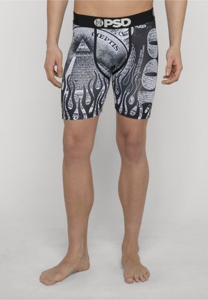 Mężczyzna ubrany w czarno-białe bokserki z wzorami banknotów dolarowych i płomieni, stojący boso na jednolitym szarym tle.