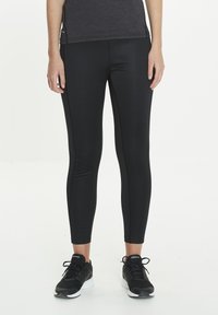 Leggings de sport noirs en tissu extensible, avec une coupe slim et des coutures latérales discrètes, assortis à des chaussures de sport noires.