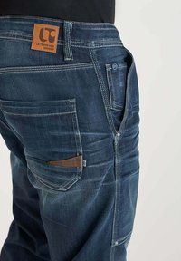 Jeans de mezclilla azul oscuro con una etiqueta de parche de cuero, costuras en contraste, bolsillos frontales y traseros, y cremallera. Detalles de desgaste visibles.